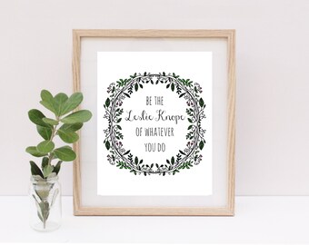 May include: A framed print with a white background and a black floral wreath with green leaves and pink berries. The text inside the wreath reads "Be the Leslie Knope of whatever you do."