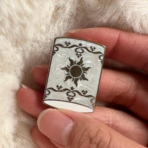 May include: A rectangular enamel pin with a gold border. The pin features a white background with a sun design in the centre. Decorative swirls and stars adorn the edges. The pin is held by a person.
