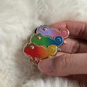 May include: A set of three colorful chameleon enamel pins. The pins are stacked on top of each other, with the top one being purple, the middle one green, and the bottom one red and orange. Each pin has a gold outline.
