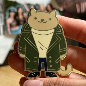 Puede incluir: Un pin de esmalte dorado con un gato de dibujos animados que lleva un abrigo verde, un su&eacute;ter color crema y pantalones azules. El gato tiene una expresi&oacute;n sonriente y zapatos negros. El pin est&aacute; en la mano de una persona.