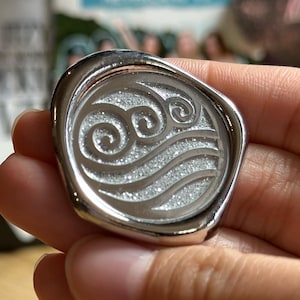 May include: A silver-colored, oval-shaped pendant with a raised border. The center features a swirling water symbol with a glittery, textured surface. The pendant is held in a hand.