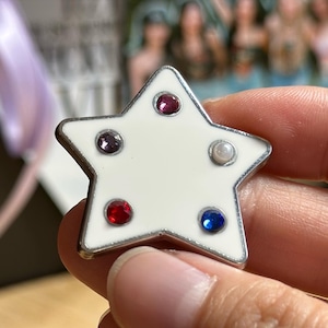 May include: A star-shaped pin with a white enamel finish and a silver-colored border. The pin is adorned with five colorful gemstones: red, purple, blue, and a pearl. The pin is held in a hand.