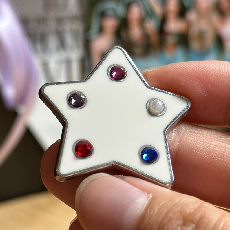May include: A star-shaped pin with a white enamel finish and a silver-colored border. The pin is adorned with five colorful gemstones: red, purple, blue, and a pearl. The pin is held in a hand.