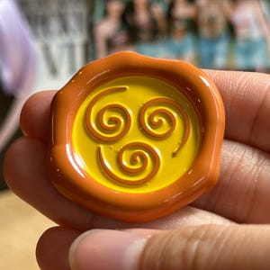 May include: A round, orange-yellow wax seal with a spiral design. The seal features a bright yellow center with three brown spiral shapes. The outer edge is a darker orange, creating a contrasting border. The design is smooth and glossy.
