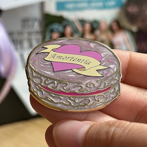 May include: A round, gold-colored enamel pin with a cake design. The pin features a pink heart and a banner with the word "Amortentia". The cake has a pink and white frosted top with small pink hearts.