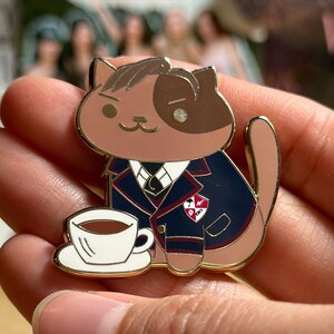 Puede incluir: Un pin de esmalte con forma de gato, en tonos marr&oacute;n y beige, que lleva una blazer azul marino con ribetes rojos. El pin incluye una taza y un platillo blancos. El gato tiene una expresi&oacute;n severa y un contorno dorado.