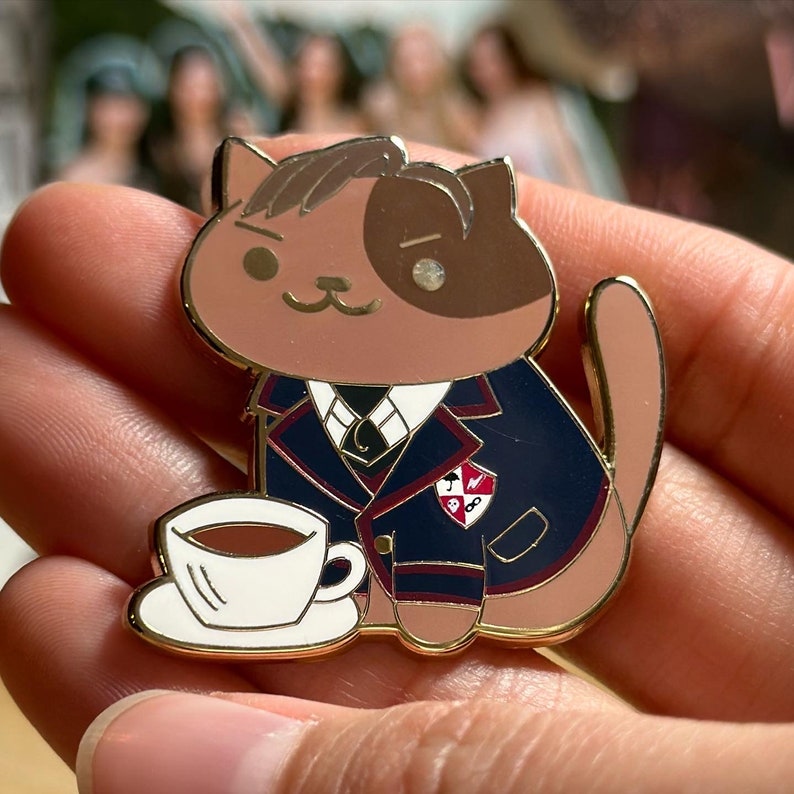 Puede incluir: Un pin de esmalte con forma de gato, en tonos marr&oacute;n y beige, que lleva una blazer azul marino con ribetes rojos. El pin incluye una taza y un platillo blancos. El gato tiene una expresi&oacute;n severa y un contorno dorado.