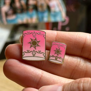 May include: Two pink enamel pins in the shape of lanterns, with a gold sun design and gold trim. The pins are in different sizes, with the larger one measuring approximately 1 inch tall. The background is blurred.