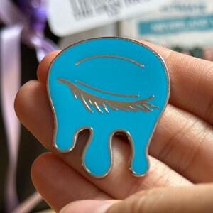 May include: A teal enamel pin with a silver outline and a closed eye design. The eye has silver eyelashes and is set against a teal background. The pin has a dripping shape.