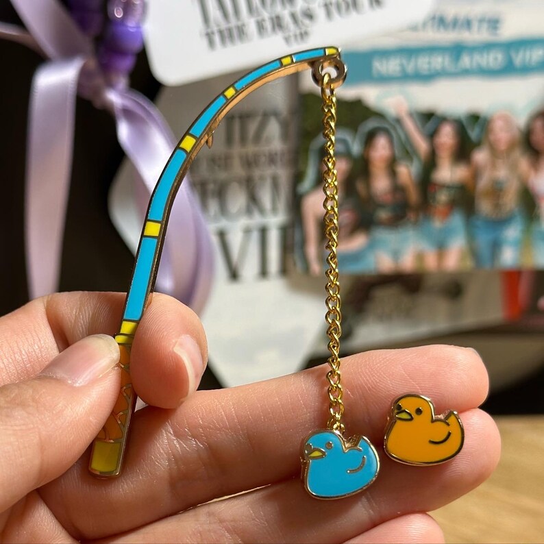 K&ouml;nnte beinhalten: Ein goldfarbener Angelruten-Pin mit blauem und gelbem Design, verbunden mit einer goldenen Kette. Die Kette f&uuml;hrt zu einem blauen Enten-Pin und einem orangefarbenen Enten-Pin. Die Pins sind wahrscheinlich Emaille.