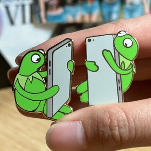 May include: Two enamel pins featuring Kermit the Frog holding a gray smartphone. The pins have a gold border and are in a cartoon style. The frog is green with a white collar and eyes.