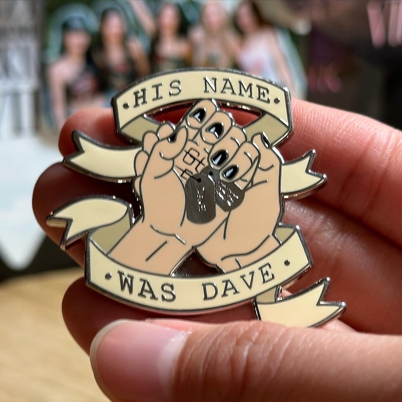 Puede incluir: Un pin met&aacute;lico con dos manos entrelazadas sosteniendo chapas de identificaci&oacute;n. El pin tiene una pancarta con el texto "HIS NAME WAS DAVE" en letras negras. Las manos son de color bronceado con esmalte de u&ntilde;as negro.