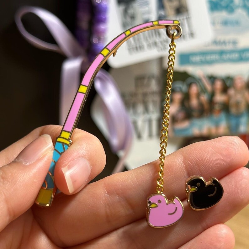 K&ouml;nnte beinhalten: Ein skurriles Emaille-Pin-Set mit einer rosa und gelben Angelrute mit einer goldenen Kette und zwei Enten-Pins. Eine Ente ist rosa, die andere ist schwarz. Die Pins sind wahrscheinlich f&uuml;r eine Jacke, einen Rucksack oder ein anderes Accessoire.