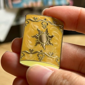 May include: A rectangular enamel pin with a yellow background and gold trim. The pin features a sun design in the center and decorative flourishes. The pin is held between fingers.