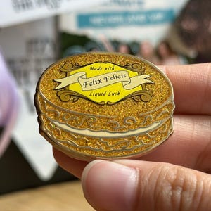 Puede incluir: Un pin de esmalte dorado con forma de pastel en capas con detalles brillantes. El pin presenta una pancarta amarilla con el texto "Made with Felix Felicis Liquid Luck".