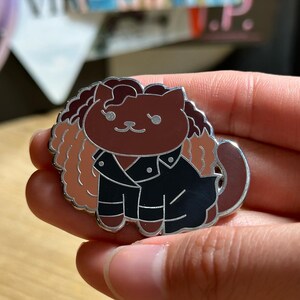 Puede incluir: Un pin de esmalte con forma de gato, con cuerpo marr&oacute;n, chaqueta negra y melena marr&oacute;n claro. El pin tiene un borde plateado y se sostiene en una mano.