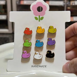 K&ouml;nnte beinhalten: Neun bunte Enten-Anstecknadeln auf einer wei&szlig;en Karte mit einer rosa Blume und dem Wort "HANDMADE". Die Anstecknadeln sind in verschiedenen Farben erh&auml;ltlich, darunter Rot, Gelb, Gr&uuml;n, Blau, Lila, Rosa, Wei&szlig; und Schwarz. Jede Nadel hat eine goldene Umrandung.