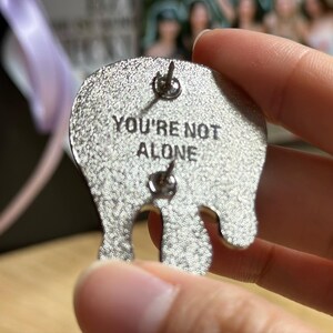 May include: Silver-colored enamel pin with the phrase "YOU'RE NOT ALONE" in black lettering. The pin has a unique, abstract shape with two pin backs for secure attachment. The pin is held in a person's hand.