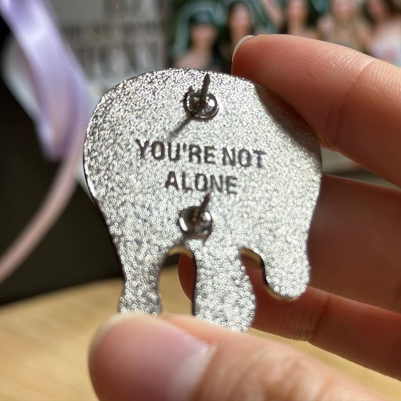 May include: Silver-colored enamel pin with the phrase "YOU'RE NOT ALONE" in black lettering. The pin has a unique, abstract shape with two pin backs for secure attachment. The pin is held in a person's hand.
