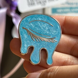 May include: A light blue enamel pin with a silver-colored outline. The pin features a stylized design of a teardrop shape with a silver feather and a curved line. The pin has a textured, marbled appearance.