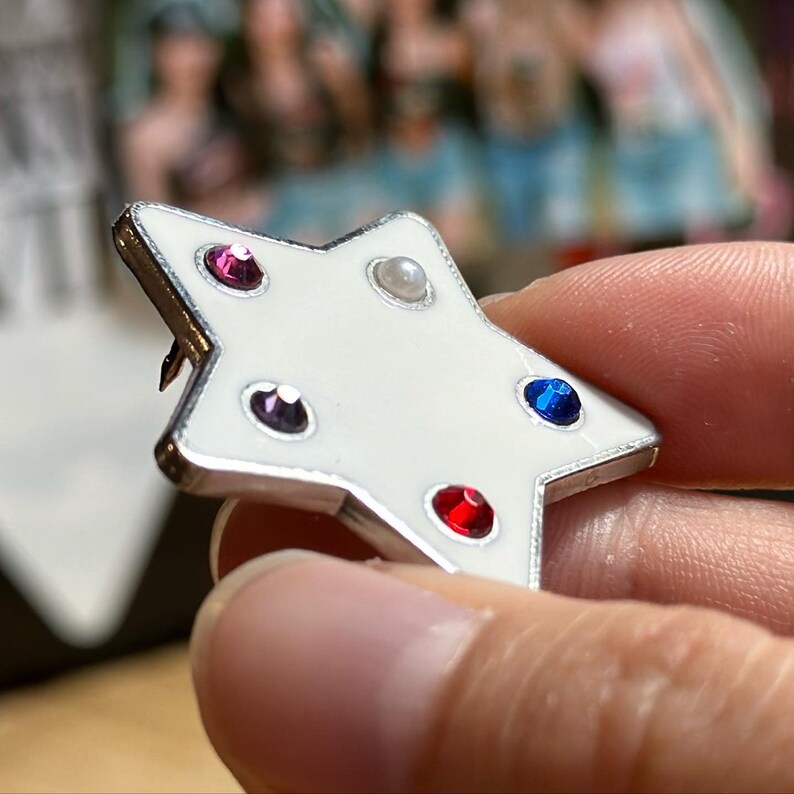 May include: A white star-shaped enamel pin with a silver border. The pin is decorated with red, blue, purple, and pearl-colored gemstones. The pin is held in a hand.