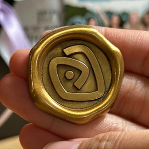 Könnte beinhalten: Ein runder, antikgoldfarbener Wachssiegel mit einem zentralen Design. Das Design zeigt eine stilisierte Herzform mit einem kleinen Kreis in der Mitte. Das Siegel hat einen erhabenen Rand und eine leicht strukturierte Oberfläche.