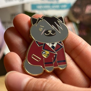 Puede incluir: Un pin dorado con un gato de dibujos animados con pelaje gris y pelo negro. El gato lleva una chaqueta burdeos con ribetes azul marino y una camisa blanca con cuello. Un peque&ntilde;o emblema est&aacute; en la chaqueta.