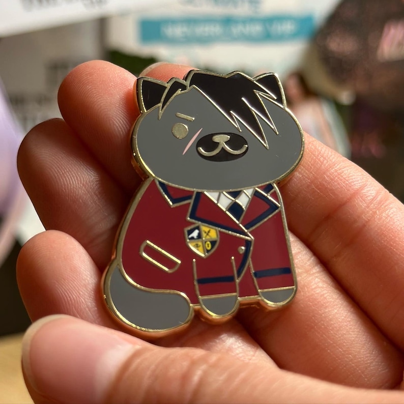 Puede incluir: Un pin dorado con un gato de dibujos animados con pelaje gris y pelo negro. El gato lleva una chaqueta burdeos con ribetes azul marino y una camisa blanca con cuello. Un peque&ntilde;o emblema est&aacute; en la chaqueta.