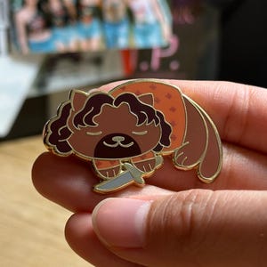 Puede incluir: Un pin de esmalte de gato marr&oacute;n con contorno dorado, sosteniendo un cuchillo plateado. El gato lleva un atuendo naranja con peque&ntilde;os estampados florales. El pin se sostiene en una mano.