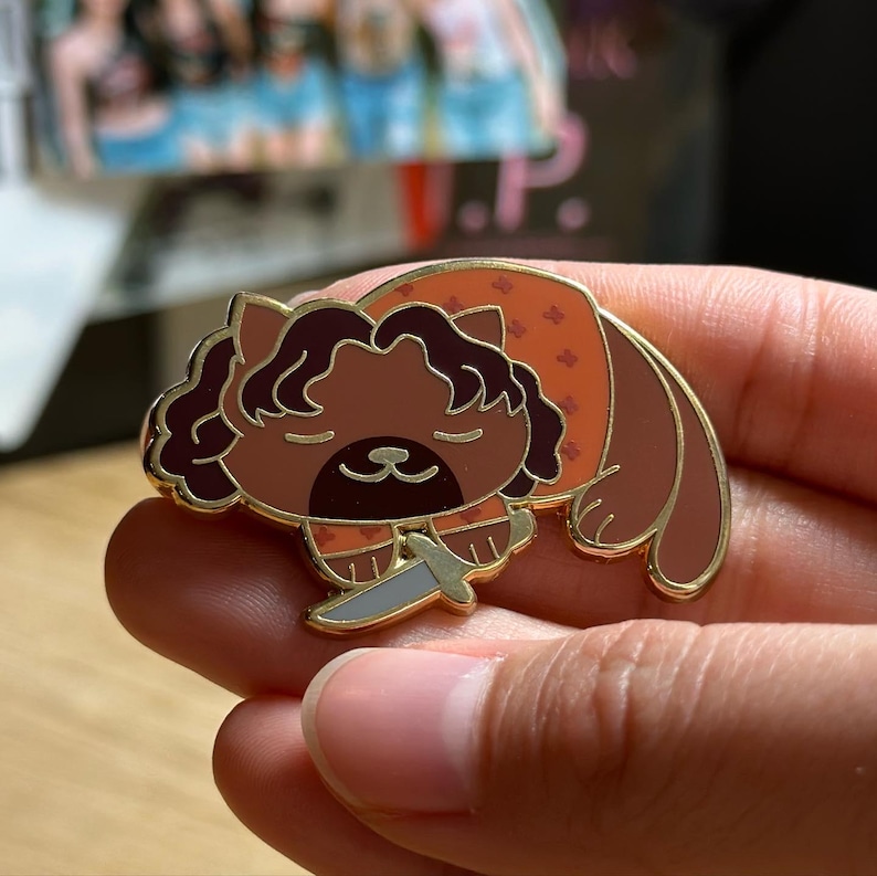 Puede incluir: Un pin de esmalte de gato marr&oacute;n con contorno dorado, sosteniendo un cuchillo plateado. El gato lleva un atuendo naranja con peque&ntilde;os estampados florales. El pin se sostiene en una mano.