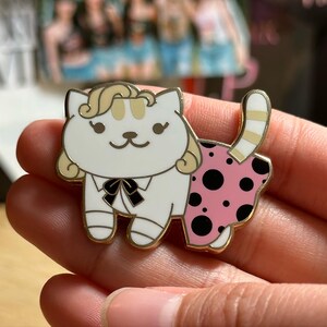 Puede incluir: Pin de esmalte de gato blanco con contorno dorado. El gato tiene pelo rubio, pajarita negra y falda rosa con lunares negros. Un accesorio lindo y caprichoso.