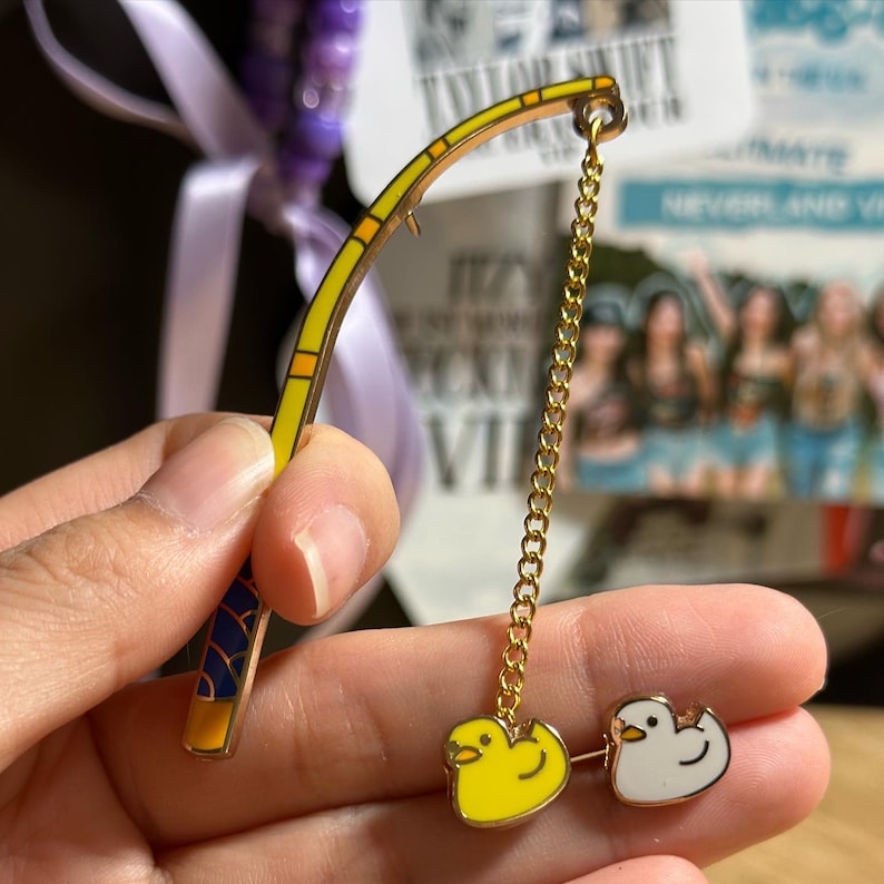 K&ouml;nnte beinhalten: Eine goldfarbene Angelruten-Anstecknadel mit gelb-blauem Griff. Eine goldene Kette h&auml;ngt an der Rute und ist mit zwei Enten-Anh&auml;ngern verbunden, einer gelben und einer wei&szlig;en. Die Nadel wird in einer Hand gehalten.