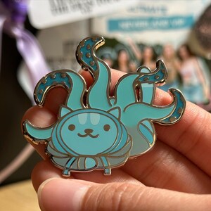 Puede incluir: Un pin de esmalte turquesa y dorado con un gato de dibujos animados con tent&aacute;culos de pulpo. El gato tiene una cara sonriente y detalles a rayas. Los tent&aacute;culos son de un turquesa m&aacute;s claro con detalles dorados.