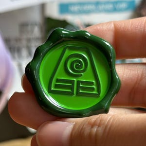 Puede incluir: Un sello de cera verde con un diseño circular que presenta una espiral y formas geométricas. El sello es de color verde vibrante y tiene un acabado brillante. El diseño está centrado en el sello.