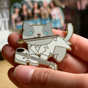 Puede incluir: Un pin de esmalte dorado que representa un gato gris y blanco tocando el viol&iacute;n. El gato lleva un traje blanco y sostiene un arco en la boca. El viol&iacute;n es blanco con detalles negros. El pin tiene un dise&ntilde;o detallado y un acabado pulido.