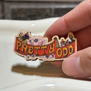 Puede incluir: Un pin de madera con las palabras "PRETTY ODD" en naranja sobre una pancarta roja. El pin está decorado con flores rosas y amarillas y una mariposa azul. El pin está sostenido por una persona.