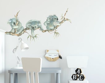 May include: A set of three koala bear wall decals, featuring a mother koala and two baby koalas, perched on a branch with green leaves. The decals are a light blue color and are perfect for adding a touch of whimsy to a child's room.