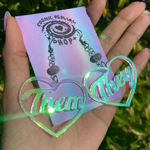 Peut inclure: Une paire de boucles d'oreilles en forme de cœur irisées avec les mots "Them" et "They" inscrits. Les boucles d'oreilles sont sur une chaîne argentée avec une petite perle violette.