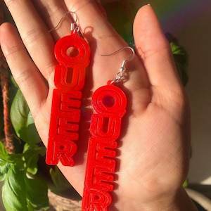 May include: A pair of red acrylic earrings with the word "QUEER" spelled out in a bold, layered font.