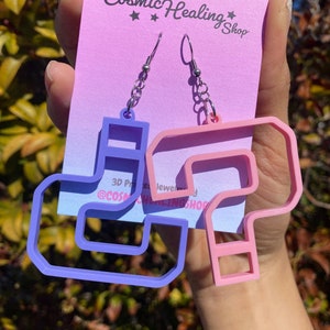 May include: A pair of earrings featuring a purple and pink question mark design. The earrings are made of 3D printed jewellery and are sold by The Cosmic Healing Shop.