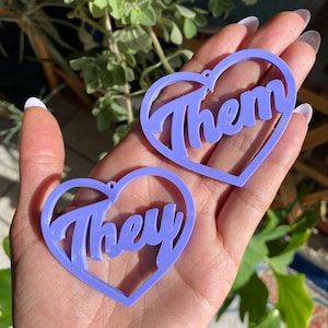 Peut inclure: Deux boucles d'oreilles en forme de cœur de couleur lavande avec les mots "They" et "Them" écrits dans une police de caractères stylisée.