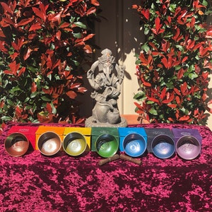 May include: Seven singing bowls in different colours, each with a unique design, are arranged in a row on a red velvet surface. The bowls are in front of a statue of Ganesha, the Hindu god of beginnings, prosperity, and good fortune. The bowls are used for sound healing and meditation.