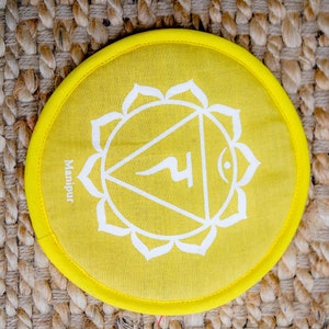 Op de afbeelding: Geel rond kussen met een wit symbool en het woord "Manipura" erop gedrukt. Het symbool is een driehoek met een cirkel bovenaan en een gebogen lijn erin. Het kussen ligt op een geweven bruin oppervlak.