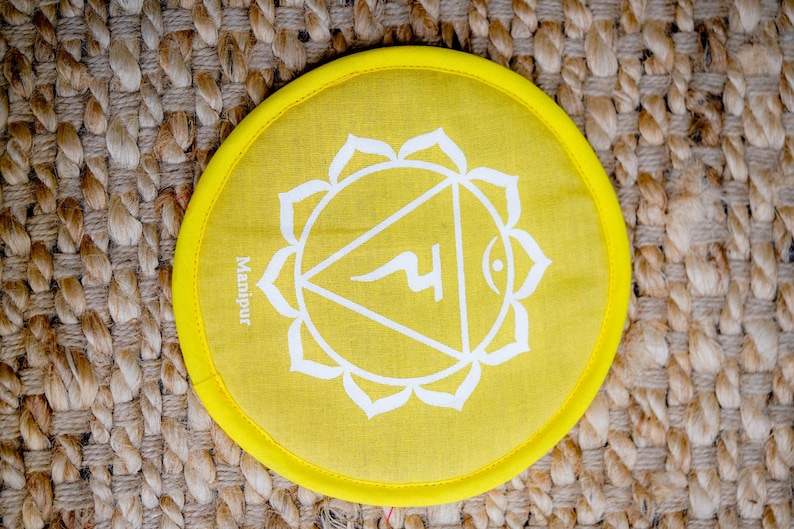 Op de afbeelding: Geel rond kussen met een wit symbool en het woord "Manipura" erop gedrukt. Het symbool is een driehoek met een cirkel bovenaan en een gebogen lijn erin. Het kussen ligt op een geweven bruin oppervlak.