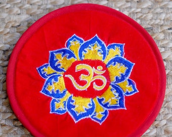 Op de afbeelding: Een rond rood kussen met een geborduurd blauw en geel lotusbloemmotief en het symbool "Om" in het midden.
