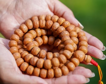 May include: A close-up of a brown wooden mala bead necklace with a red and yellow tassel. The mala beads are held in the palm of a hand.