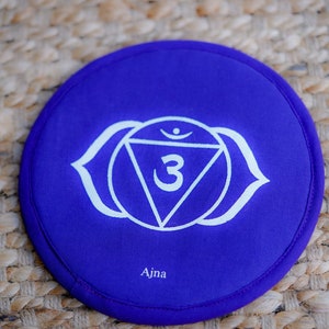 Op de afbeelding: Rond paars kussen met een wit symbool van de derde oog chakra, Ajna, met het nummer 3 in een driehoek.