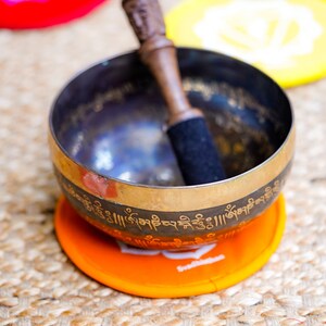 Op de afbeelding: Een zangkom met een houten slaagstok, rustend op een oranje kussen met een wit lotusontwerp. De kom is versierd met gouden letters en staat op een geweven oppervlak.