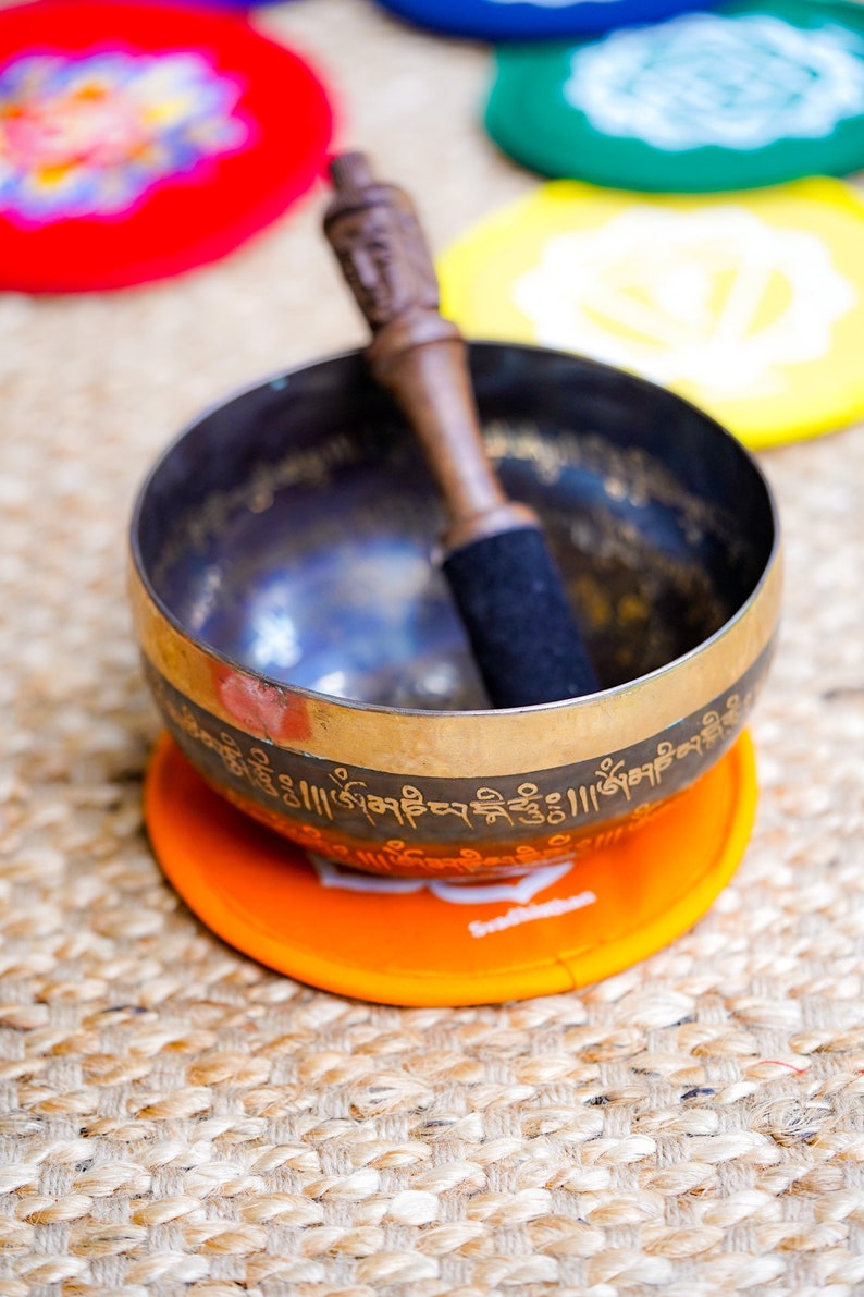 Op de afbeelding: Een zangkom met een houten slaagstok, rustend op een oranje kussen met een wit lotusontwerp. De kom is versierd met gouden letters en staat op een geweven oppervlak.