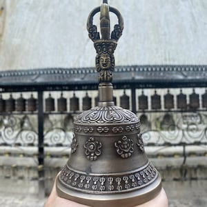 May include: A bronze bell with intricate carvings and a decorative handle. The bell has a floral design and a small face on the handle.